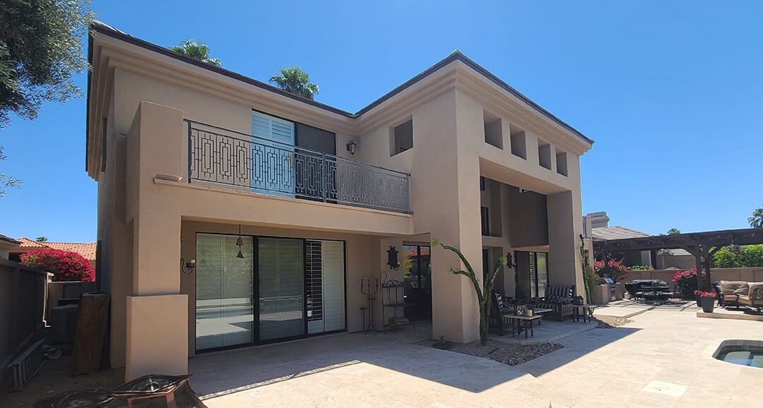 Before painting: two-story tan stucco home with a second-floor balcony and stone patio